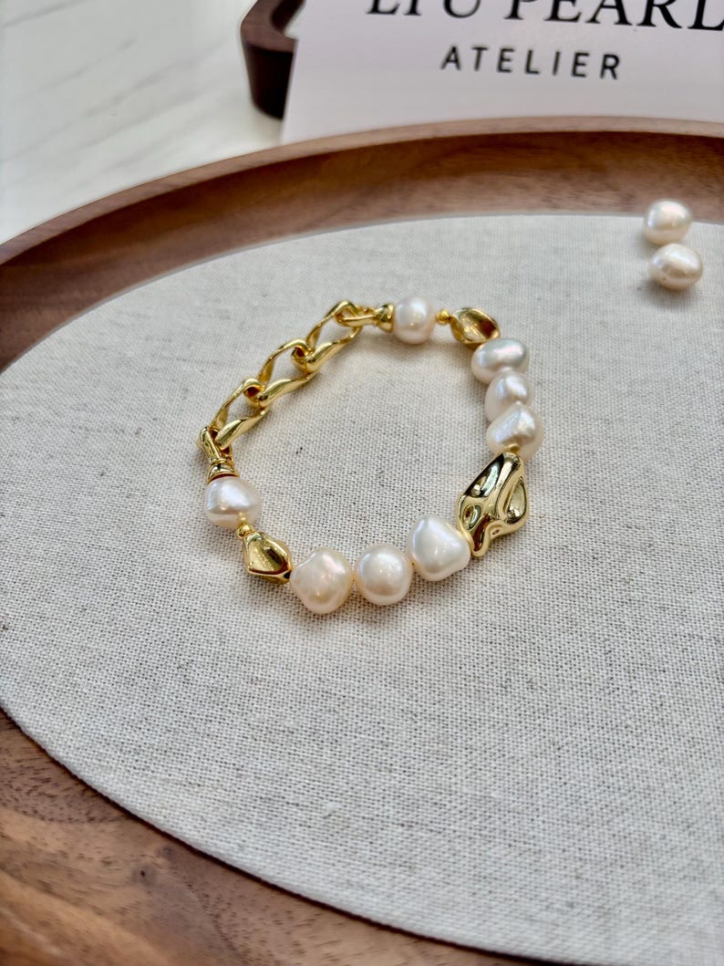 May include: A gold-toned bracelet featuring a combination of chain links and organic-shaped pearls. The bracelet has a mix of textures, with the smooth pearls contrasting the textured metal. The bracelet is displayed on a neutral linen surface.