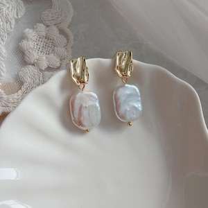 May include: Gold-coloured earrings with a textured, rectangular top and a large, irregular, white pearl drop. The earrings are displayed on a white, scalloped dish.