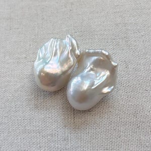May include: Two irregularly shaped, lustrous white pearls with a pearlescent sheen. The pearls have a unique, organic form, reflecting light and displaying subtle colour variations. They rest on a textured, neutral-toned fabric.