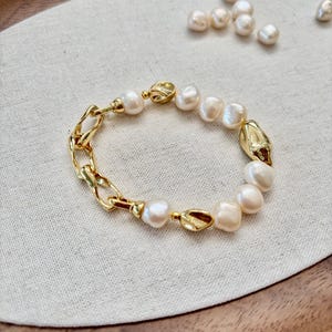May include: A gold-toned bracelet featuring a combination of freshwater pearls and chain links. The bracelet has a mix of smooth and textured elements, with the pearls varying in size and shape. The clasp and connecting pieces are also gold-toned.