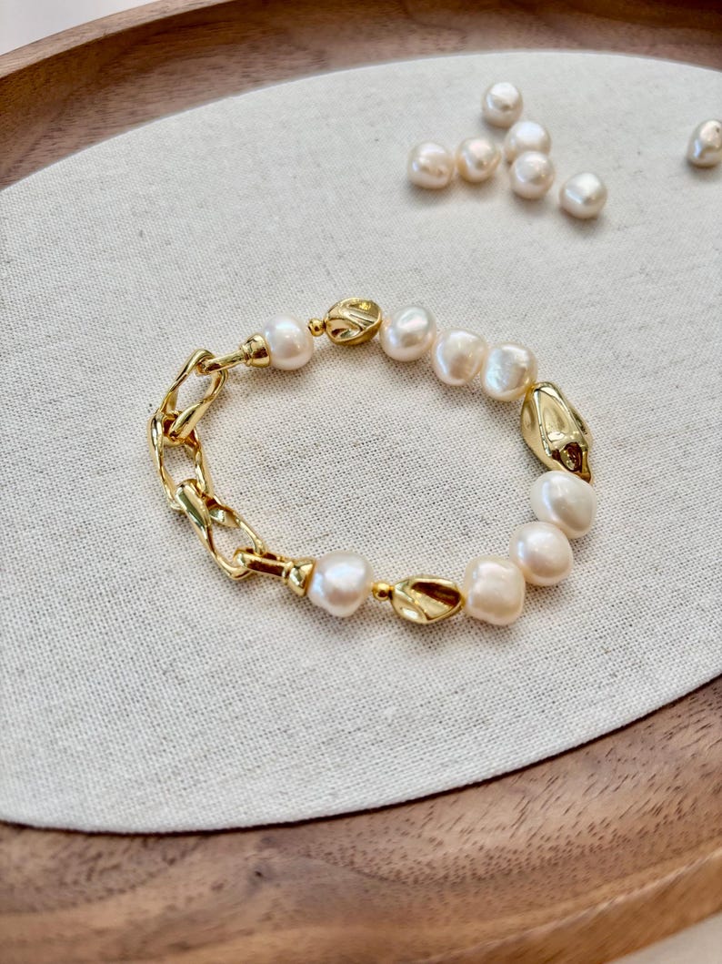 May include: A gold-toned bracelet featuring a combination of freshwater pearls and chain links. The bracelet has a mix of smooth and textured elements, with the pearls varying in size and shape. The clasp and connecting pieces are also gold-toned.