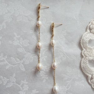 May include: A pair of elegant gold-coloured earrings. Each earring features three off-white pearls suspended from a fine chain. The top of each earring has a unique, organic shape. The earrings are displayed against a soft, floral-patterned background.