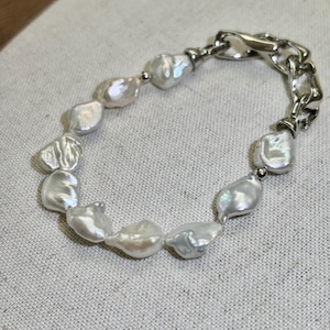 May include: A white pearl bracelet with a silver chain clasp. The bracelet features irregularly shaped, lustrous white pearls strung together. The silver chain adds a modern contrast to the classic pearl design. The bracelet is displayed on a neutral fabric background.