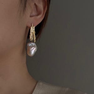 May include: A gold-toned earring featuring a textured, irregular metal top piece with a rough, organic finish. Suspended from this metallic element is a large, lustrous baroque pearl with an irregular, bulbous shape and a soft, iridescent sheen.