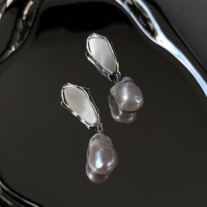 May include: A pair of elegant earrings with a silver-toned, irregular frame and a pearlescent insert, each suspending a large, teardrop-shaped pearl. The earrings are on a reflective, dark surface.