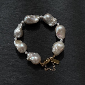May include: A bracelet featuring large, irregularly shaped, off-white pearls. Smaller, round pearls are interspersed between the larger ones. The bracelet is secured with a gold-colored clasp and a small tag.