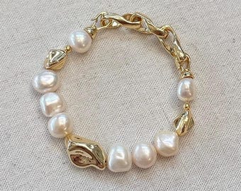 Peut inclure: Un bracelet doré composé d'un mélange de grosses perles irrégulières et de maillons dorés texturés. Le bracelet est doté d'un fermoir mousqueton. Les perles sont blanches avec une légère teinte rosée. Les maillons dorés ont un design organique.