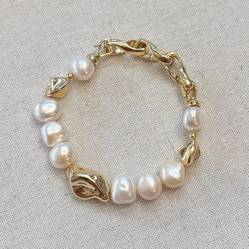 May include: A gold-toned bracelet featuring a mix of large, irregular pearls and textured gold links. The bracelet has a lobster clasp closure. The pearls are white with a slight pink hue. The gold links have an organic, flowing design.