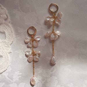 Op de afbeelding: Een paar goudkleurige oorbellen. Elke oorbel heeft een cluster van crèmekleurige, bloemblad-vormige parels die aan een kleine ring zijn bevestigd. Een delicate gouden ketting strekt zich uit vanaf de cluster en eindigt in een enkele, grotere parel. De oorbellen zijn geplaatst tegen een zachte, patroonachtergrond.