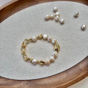May include: A gold-toned bracelet featuring a combination of irregular pearls and chain links. The bracelet is displayed on a neutral linen surface within a wooden tray, with additional pearls scattered nearby.