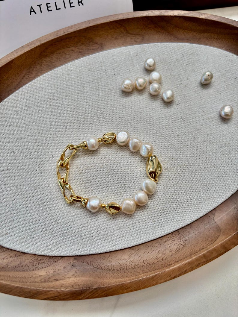 May include: A gold-toned bracelet featuring a combination of irregular pearls and chain links. The bracelet is displayed on a neutral linen surface within a wooden tray, with additional pearls scattered nearby.