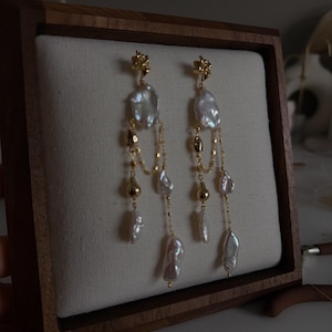 May include: A pair of gold-tone earrings with irregular white pearls and small gold beads. The earrings have a floral design at the top and dangle with multiple strands of pearls and beads, displayed in a wooden jewelry box.