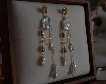 May include: A pair of gold-tone drop earrings featuring irregular white baroque pearls and delicate gold chains. Each earring has a textured gold stud top, multiple dangling pearl segments of varying shapes, and small gold beads suspended on thin chains.