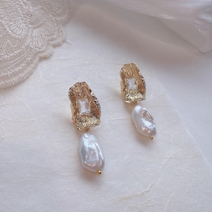 May include: A pair of gold-toned earrings featuring a textured, irregular metal setting with a central clear rectangular stone. Each earring is finished with a dangling, white, organic-shaped baroque pearl attached to the base of the metal piece.
