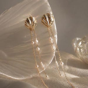 May include: Gold seashell earrings with dangling pearl details. The earrings have a detailed seashell design at the top, with delicate chains and strands of pearls hanging down. The earrings are set against a soft, neutral background.