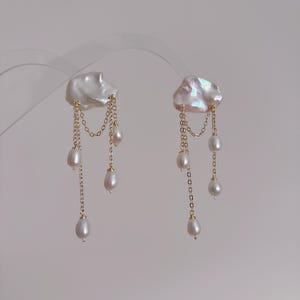 May include: A pair of gold-tone chain earrings featuring large, irregular white baroque pearls at the top. Each earring has three delicate gold chains hanging from the pearl, with smaller teardrop-shaped white pearls attached to the ends of the chains.