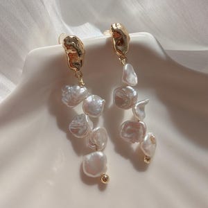 May include: Drop earrings with irregular, off-white pearls hanging from textured gold-coloured metal studs. The earrings are presented on a white, shell-shaped surface, with soft lighting enhancing their lustre.