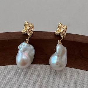 May include: A pair of gold earrings with textured tops and large, pearlescent white baroque pearls. The earrings hang from delicate gold connectors, showcasing a unique design. The earrings are displayed on a wooden surface. The pearls have a beautiful iridescent sheen.