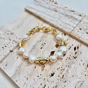 May include: A gold-toned bracelet featuring irregular white pearls and a chunky chain clasp. The bracelet has gold connectors between the pearls. The jewelry is displayed on a textured, light-colored surface.