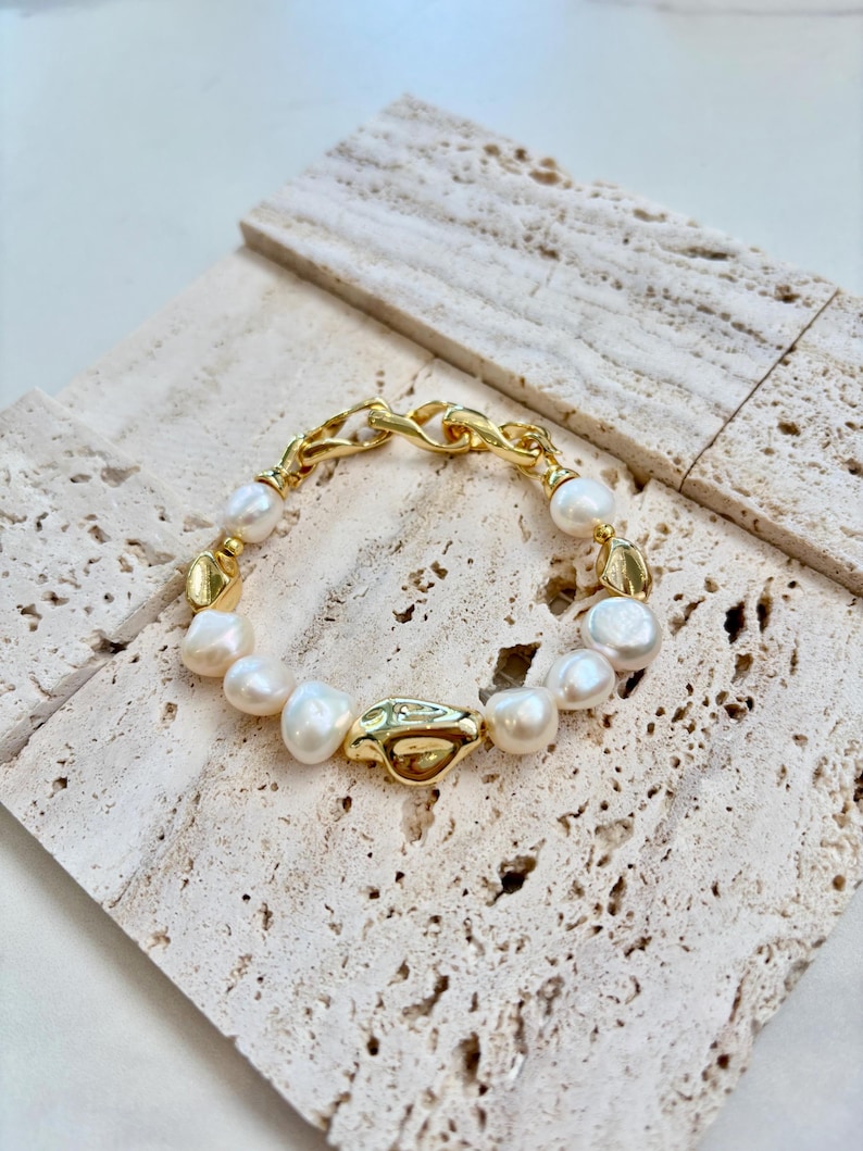 May include: A gold-toned bracelet featuring irregular white pearls and a chunky chain clasp. The bracelet has gold connectors between the pearls. The jewelry is displayed on a textured, light-colored surface.