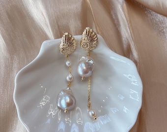 May include: A pair of gold-toned earrings with a seashell design at the top. Each earring features a large, irregular pearl suspended from a delicate chain, along with smaller pearls. The earrings are displayed on a white, shell-shaped dish.