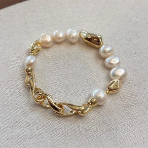 May include: A gold-toned bracelet featuring a mix of large, irregular pearls and textured gold links. The pearls vary in size and have a creamy white color. The bracelet is set against a neutral background.