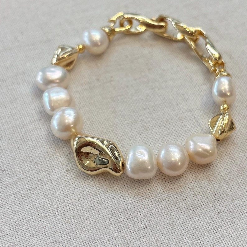 May include: A gold-toned bracelet featuring a mix of large, irregular pearls and textured gold links. The bracelet has a unique design with a combination of organic shapes and classic elements.