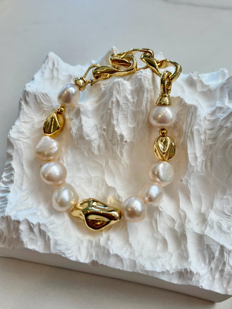 May include: A gold-toned bracelet featuring a chain link design and a series of freshwater pearls. The bracelet also includes gold-colored, irregularly shaped beads. The bracelet is displayed on a textured white surface.