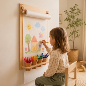 Op de afbeelding: Een aan de muur gemonteerde papierrolhouder van licht hout met een plankje met vier kleine bekers gevuld met kleurpotloden. Een lang vel wit papier hangt naar beneden, met een kleurrijke handgetekende illustratie van een huis, zon, wolken, boom en bloemen.
