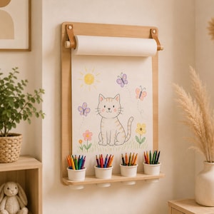 May include: A wall-mounted light wood paper roll holder featuring a drawing of a cat, sun, flowers, and butterflies on white paper. The unit includes a bottom shelf with four white cups holding colored pencils for drawing and creative activities.