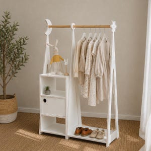 May include: A white wooden clothing rack featuring a moon and star design on the side supports, a natural wood hanging rod, three storage shelves, and a closed cabinet. The rack holds several light-colored garments on hangers and a plush rabbit toy.