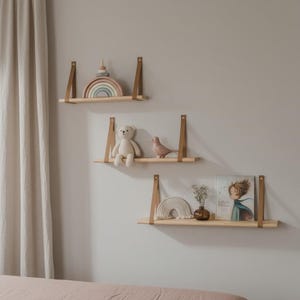 May include: Three wooden shelves with leather straps on a light grey wall. The shelves display a wooden rainbow, a teddy bear, a bird figurine, a book, and other decorative items. The shelves are made of light-coloured wood.