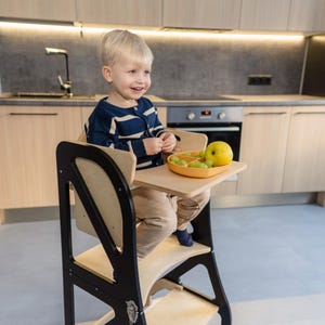 May include: A wooden learning tower for toddlers, featuring a black frame and natural wood elements. The tower has a small tray with a yellow divided plate holding grapes and an apple. A young child is seated in the tower, smiling.