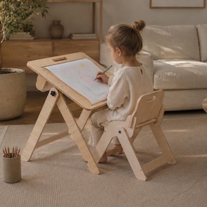 Può includere: Un tavolo da disegno in legno chiaro con superficie inclinabile regolabile e una sedia in legno abbinata. Il tavolo sostiene un foglio di carta bianca con un disegno e un contenitore cilindrico pieno di matite si trova sul pavimento vicino.