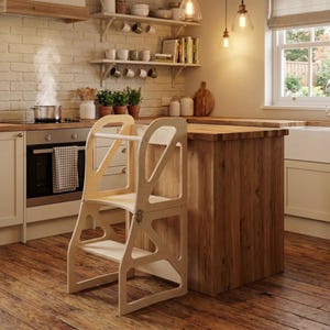 May include: A light-coloured wooden learning tower with a tall frame, a safety rail, and a step platform. The structure features cut-out side panels and a hinge mechanism, designed to provide an elevated standing surface for children in a kitchen.