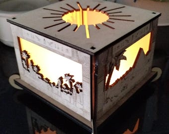 May include: A wooden, laser-cut light box with an Egyptian theme. The box features cut-out designs of palm trees, pyramids, and hieroglyphs. The top has a sunburst design. The interior glows with a warm, yellow light, creating a silhouette effect.