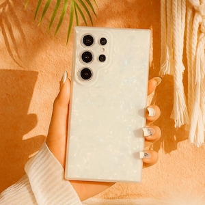 Op de afbeelding: Een witte parelmoer telefoonhoes met een cameramodule met meerdere zwarte lenzen. De hoes wordt in een hand gehouden, met een zachte, getextureerde achtergrond. De hoes heeft een rechthoekige vorm.