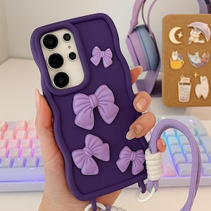May include: Purple phone case with raised lavender bows. The case has a matching lavender wrist strap with white accents. The phone case is held in a hand.