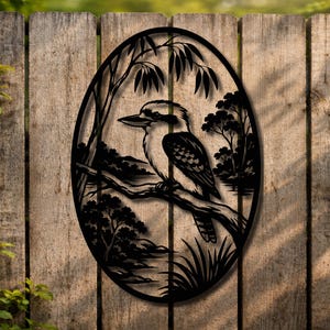 May include: Black metal wall art depicting a kookaburra bird on a branch, set within an oval frame. The artwork includes trees and a landscape, ideal for garden or interior decoration.