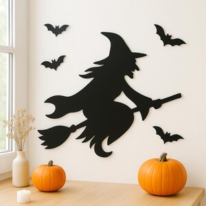 May include: Black witch silhouette on a broomstick with several bat cutouts on a white wall. Two orange pumpkins and a candle sit on a wooden surface. A vase with dried flowers is also present, creating a Halloween theme.