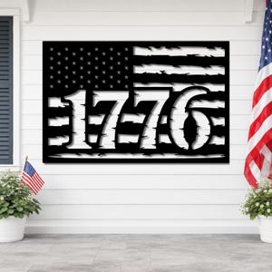 May include: Black metal wall art depicting the American flag with the year "1776" cut out. The flag design features stars in the upper left corner and stripes. The artwork is displayed outdoors, with a small American flag and potted plants visible.
