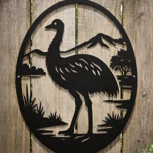 May include: Black metal wall art depicting an emu within an oval frame. The design includes a landscape with mountains, trees, and water. The artwork is mounted on a wooden fence, creating a rustic aesthetic.