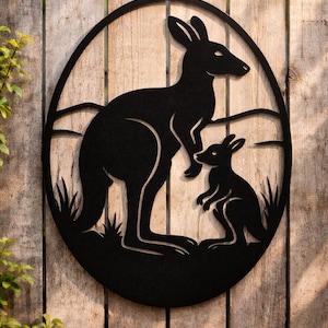 May include: Black metal wall art depicting a kangaroo and joey silhouette within an oval frame. The design includes grass and a simple landscape, mounted on a wooden fence. A decorative piece for home or garden.