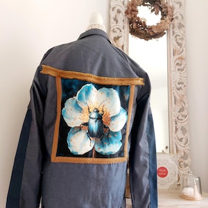 Peut inclure: Veste en jean avec une grande peinture encadrée sur le dos. La peinture représente un scarabée bleu sur une fleur blanche et bleue. La veste a des accents bleu foncé sur les manches et une bordure dorée.