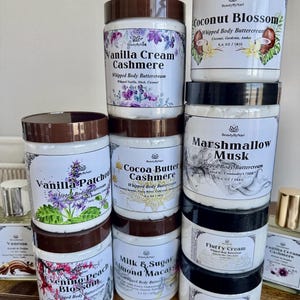 May include: A collection of whipped body buttercreams in various scents, displayed in white jars with brown lids. The labels feature floral designs and text such as "Vanilla Cream Cashmere" and "Coconut Blossom".