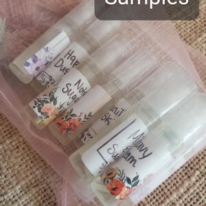 May include: A collection of small, clear sample bottles with white labels and floral designs, held in a sheer pink bag. The labels feature handwritten text, including the word "Samples" at the top.