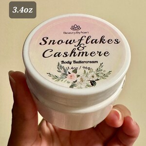 May include: A white jar of body buttercream with a floral design and the text "Snowflakes & Cashmere." The label also includes the text "Body Buttercream" and "3.4oz / 96g."