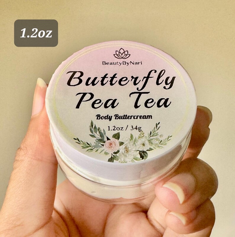 May include: A jar of body buttercream with a floral design and the text "Butterfly Pea Tea". The jar is labeled "1.2oz / 34g". The lid is light pink with a white rim. The jar is held in a hand.