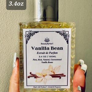May include: A rectangular glass perfume bottle with a silver cap, labeled "Vanilla Bean Extrait de Parfum." The label includes the text "3.4 OZ / 100ML" and features vanilla bean illustrations. The bottle is held by a person.