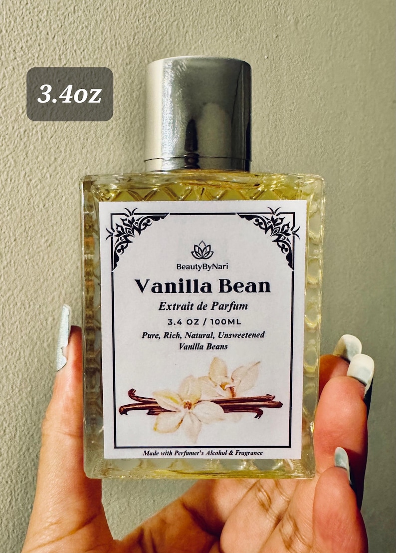 May include: A rectangular glass perfume bottle with a silver cap, labeled "Vanilla Bean Extrait de Parfum." The label includes the text "3.4 OZ / 100ML" and features vanilla bean illustrations. The bottle is held by a person.